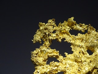 A piece of natural gold nugget with a detailed, intricate texture against a dark background. The nugget has a bright, shiny surface with various small crevices and folds.