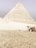 A large pyramid stands prominently, constructed with a series of layered stones in a sandy desert environment. A camel, adorned with colorful fabric, is situated to the right, with its handler standing nearby in traditional attire.