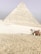 A large pyramid stands prominently, constructed with a series of layered stones in a sandy desert environment. A camel, adorned with colorful fabric, is situated to the right, with its handler standing nearby in traditional attire.