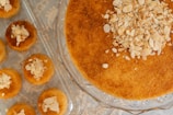 A large round pastry with a golden brown texture topped with chopped nuts is displayed on a glass cake stand. Around it, smaller pastries are arranged on a tray, also topped with nuts.