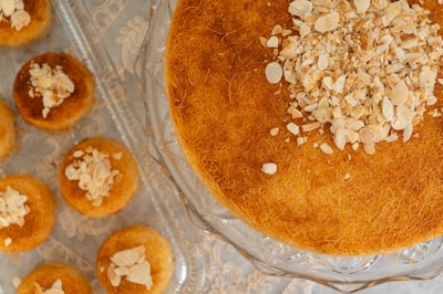 A large round pastry with a golden brown texture topped with chopped nuts is displayed on a glass cake stand. Around it, smaller pastries are arranged on a tray, also topped with nuts.
