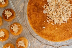 A large round pastry with a golden brown texture topped with chopped nuts is displayed on a glass cake stand. Around it, smaller pastries are arranged on a tray, also topped with nuts.