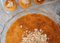 A close-up view of a dessert with a layer of finely shredded pastry, topped with sliced almonds. The dish is round and sits on a glass serving plate. In the background, there are smaller individual servings of the same dessert on a rectangular glass tray.