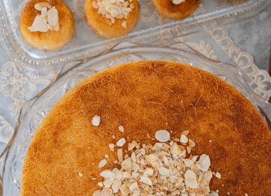 A close-up view of a dessert with a layer of finely shredded pastry, topped with sliced almonds. The dish is round and sits on a glass serving plate. In the background, there are smaller individual servings of the same dessert on a rectangular glass tray.
