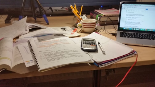 An organized yet cluttered study desk featuring an open laptop displaying a webpage, a spiral notebook with handwritten notes, a calculator, a pen, and a coffee cup. Additional study materials, books, and folders are spread across the desk, along with a jar of pencils and stationery. A monitor in the background adds to the academic setting.