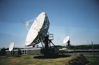 Large satellite dishes are positioned on a grassy area near a building, with a clear blue sky in the background. One prominent dish in the foreground is mounted on a metal frame, while others are visible in the distance. A rocky mound is situated nearby, and a vehicle is parked along a road in the background.