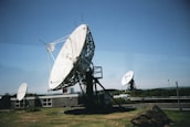 Large satellite dishes are positioned on a grassy area near a building, with a clear blue sky in the background. One prominent dish in the foreground is mounted on a metal frame, while others are visible in the distance. A rocky mound is situated nearby, and a vehicle is parked along a road in the background.
