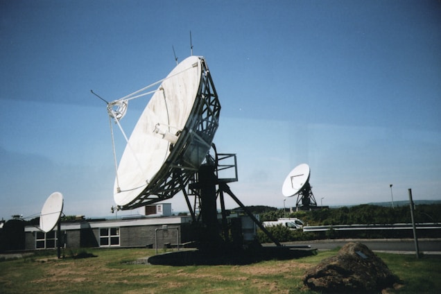 Large satellite dishes are positioned on a grassy area near a building, with a clear blue sky in the background. One prominent dish in the foreground is mounted on a metal frame, while others are visible in the distance. A rocky mound is situated nearby, and a vehicle is parked along a road in the background.