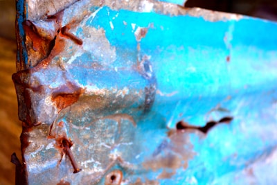 A close-up of a corroded metal sheet with a combination of blue paint and reddish-brown rust. The texture is rough with visible patches of rust along the edges and surface. The contrast between the vibrant blue and the earthy rust tones is striking.