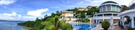 A panoramic view of a luxurious resort featuring vibrant tropical greenery surrounding a curved swimming pool. Multiple villas with large windows and balconies are situated on a lush hill. The sky is partly cloudy and the calm ocean can be seen on the left.