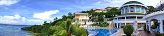 A panoramic view of a luxurious resort featuring vibrant tropical greenery surrounding a curved swimming pool. Multiple villas with large windows and balconies are situated on a lush hill. The sky is partly cloudy and the calm ocean can be seen on the left.