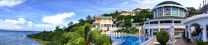 A panoramic view of a luxurious resort featuring vibrant tropical greenery surrounding a curved swimming pool. Multiple villas with large windows and balconies are situated on a lush hill. The sky is partly cloudy and the calm ocean can be seen on the left.