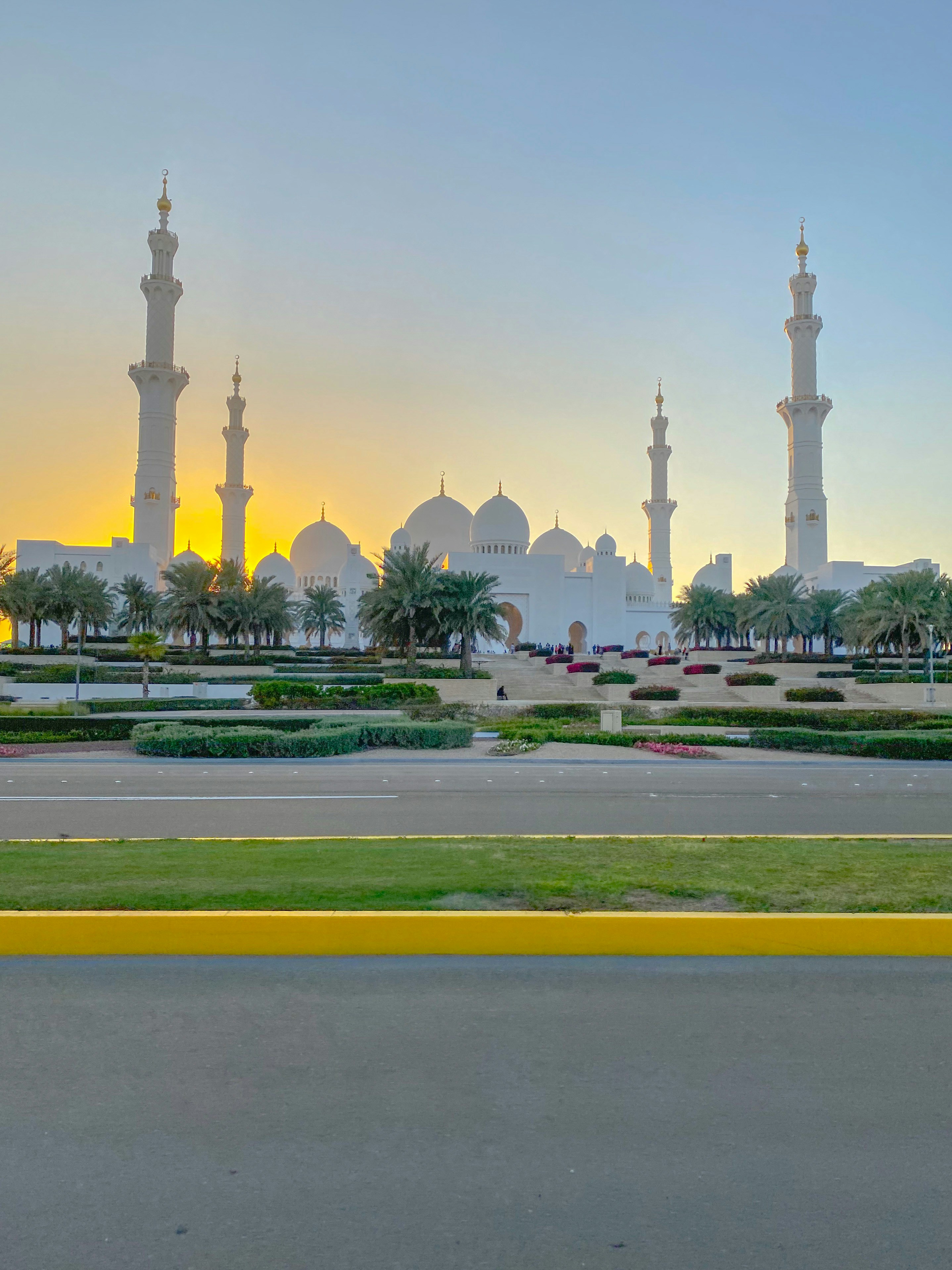 Grand mosque with white minarets and golden dome reflecting sunset light, palm trees framing the entrance in warm golden hour