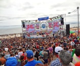 A large crowd gathers in front of a stage near the beach. The stage is decorated with colorful banners and screens displaying vibrant graphics. People appear to be enjoying an outdoor event, with many facing the large stage. The beach and ocean are visible in the background under an overcast sky.