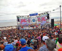 A large crowd gathers in front of a stage near the beach. The stage is decorated with colorful banners and screens displaying vibrant graphics. People appear to be enjoying an outdoor event, with many facing the large stage. The beach and ocean are visible in the background under an overcast sky.