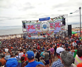 A large crowd gathers in front of a stage near the beach. The stage is decorated with colorful banners and screens displaying vibrant graphics. People appear to be enjoying an outdoor event, with many facing the large stage. The beach and ocean are visible in the background under an overcast sky.