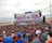 A large crowd gathers in front of a stage near the beach. The stage is decorated with colorful banners and screens displaying vibrant graphics. People appear to be enjoying an outdoor event, with many facing the large stage. The beach and ocean are visible in the background under an overcast sky.