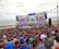 A large crowd gathers in front of a stage near the beach. The stage is decorated with colorful banners and screens displaying vibrant graphics. People appear to be enjoying an outdoor event, with many facing the large stage. The beach and ocean are visible in the background under an overcast sky.