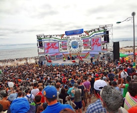 A large crowd gathers in front of a stage near the beach. The stage is decorated with colorful banners and screens displaying vibrant graphics. People appear to be enjoying an outdoor event, with many facing the large stage. The beach and ocean are visible in the background under an overcast sky.