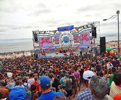 A large crowd gathers in front of a stage near the beach. The stage is decorated with colorful banners and screens displaying vibrant graphics. People appear to be enjoying an outdoor event, with many facing the large stage. The beach and ocean are visible in the background under an overcast sky.