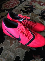 A pair of bright red athletic shoes with black accents and laces are placed on a patterned carpet. The shoes have a sleek, modern design with textured detailing and branding visible on the sides.