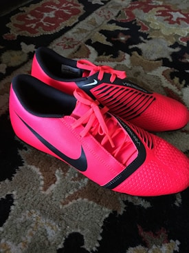 A pair of bright red athletic shoes with black accents and laces are placed on a patterned carpet. The shoes have a sleek, modern design with textured detailing and branding visible on the sides.