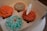 A box containing four cupcakes with intricate icing designs resembling flowers and swirls in colors of orange and teal. One cupcake has a white butterfly decoration perched on top.