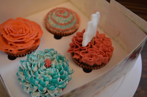 A box containing four cupcakes with intricate icing designs resembling flowers and swirls in colors of orange and teal. One cupcake has a white butterfly decoration perched on top.