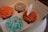 A box containing four cupcakes with intricate icing designs resembling flowers and swirls in colors of orange and teal. One cupcake has a white butterfly decoration perched on top.