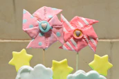 Two decorative pinwheels are fashioned in pastel shades, one with pink and blue polka dots and the other with pink and white stripes. They are attached to sticks. Below them are star and cloud-shaped objects in soft yellow and white colors. The background is a neutral beige.