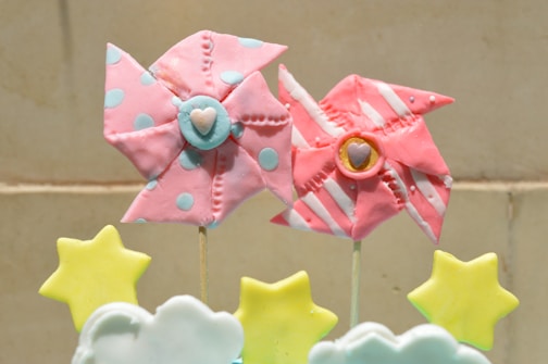 Two decorative pinwheels are fashioned in pastel shades, one with pink and blue polka dots and the other with pink and white stripes. They are attached to sticks. Below them are star and cloud-shaped objects in soft yellow and white colors. The background is a neutral beige.