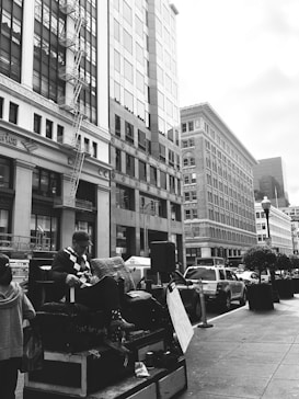 An urban street scene with towering buildings lining the sidewalk. A man is seated on a raised platform, appearing to engage in an activity with various items placed around him. Several vehicles, including taxis, are parked along the street, and a lamppost stands nearby. The atmosphere is busy yet organized.