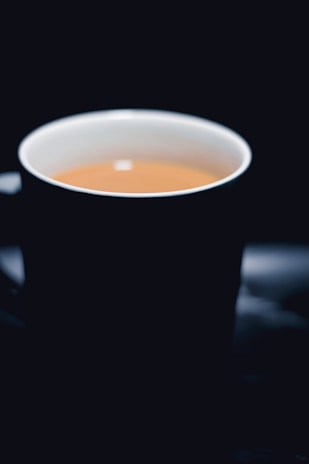 A steaming cup of tea or coffee is centered against a dark background. The cup has a light-colored interior contrasting with its darker exterior, and the warm liquid creates a cozy ambiance.