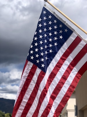 An American flag is waving against a backdrop of dark and cloudy skies. The flag's stripes and stars are prominently displayed, capturing a sense of movement and patriotism.