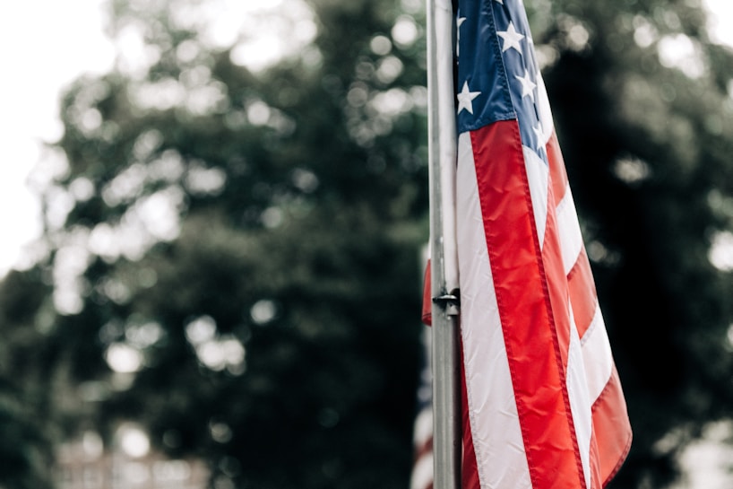 An American flag is prominently displayed, hanging from a flagpole with its red, white, and blue colors on full display. The background is blurred, showing green foliage and suggesting an outdoor setting.