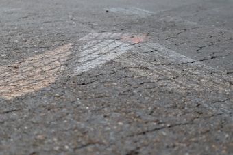 An asphalt road surface features visible cracks with a faded arrow painted on it. The arrow points in a particular direction, and the wear of the paint suggests it has been there for some time.