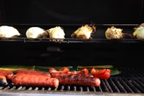 A grill loaded with various foods. On the top rack, several heads of corn are being roasted in their husks. Below, sausages are grilling alongside slices of red bell pepper and some green vegetables, possibly zucchini. The food is slightly charred, suggesting it's nearly cooked.