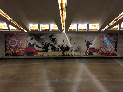 A mural depicting Soviet soldiers in battle, with symbolic imagery such as the hammer and sickle emblem and another military medal-like emblem. The scene includes triumphant soldiers, fireworks, and large Cyrillic text at the bottom. The setting is an underground space with structured ceilings and illuminated panels.