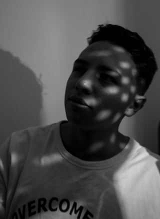 A black and white portrait of a person wearing a shirt with the word 'OVERCOME' partially visible. Patterns of light and shadow play across the face and wall, creating a dappled effect.