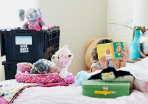 A child is sitting on a bed, holding a green children's book with characters on the cover. The bed is decorated with various stuffed animals, including a penguin, a bear, and an elephant. On the bed is a pink cushion patterned with hearts. There is a green suitcase near the child and a black box labeled 'Hive Boxx' in the background. A blue vase and other decorative items are visible on a side table.