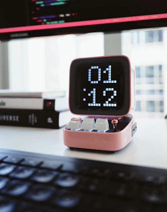 A digital clock with a retro design displays the time '01:12'. The clock has a pink casing and is placed on a desk in front of a blurred computer keyboard. Behind the clock, there are few stacked books and a computer monitor showing lines of code.
