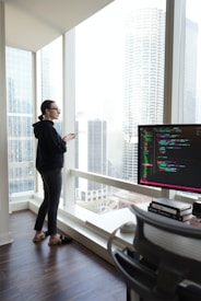 A person is standing by a large window in a modern office with a cityscape view. They are wearing a dark hoodie and looking attentively outside, holding a smartphone. A desk with a computer monitor displaying colorful code is situated in the foreground.