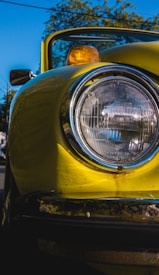 Close-up view of a vintage yellow car's headlight against a clear blue sky. The chrome detailing around the headlight is prominent, and the classic amber side marker light is also visible. The reflection on the car's surface adds an element of shine.