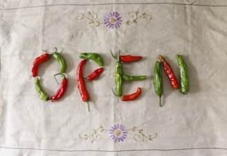 Chili peppers are arranged on a cloth to spell the word 'OPEN', with a display of alternating red and green peppers. The background features a fabric with a checkered texture and small embroidered flowers in purple and yellow.