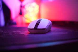 A wireless computer mouse is placed on a surface with a soft-focus background featuring colorful lights, creating a vibrant and modern atmosphere. The sharp focus on the mouse contrasts with the blurred, glowing bokeh lights in shades of pink, purple, and yellow.