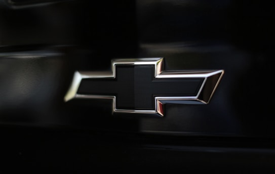 A close-up view of a metallic emblem shaped like a cross with rounded edges, placed against a dark background.