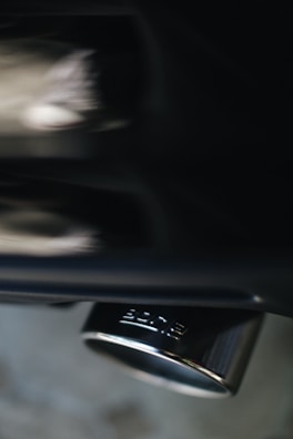 A close-up of a metallic exhaust pipe with a shiny surface, reflecting light and showing the brand logo imprinted on it. The background appears blurred, emphasizing the focus on the exhaust.