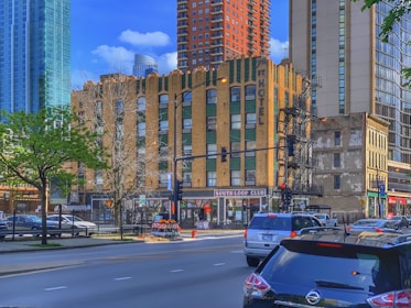 A bustling urban scene with a mix of modern and older architecture. A brick building with green accents and large '7th St Hotel' signage occupies the center. Nearby, taller modern buildings are visible. Trees with fresh green leaves line the street alongside parked cars and moving traffic.