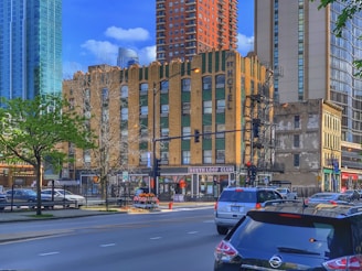 A bustling urban scene with a mix of modern and older architecture. A brick building with green accents and large '7th St Hotel' signage occupies the center. Nearby, taller modern buildings are visible. Trees with fresh green leaves line the street alongside parked cars and moving traffic.