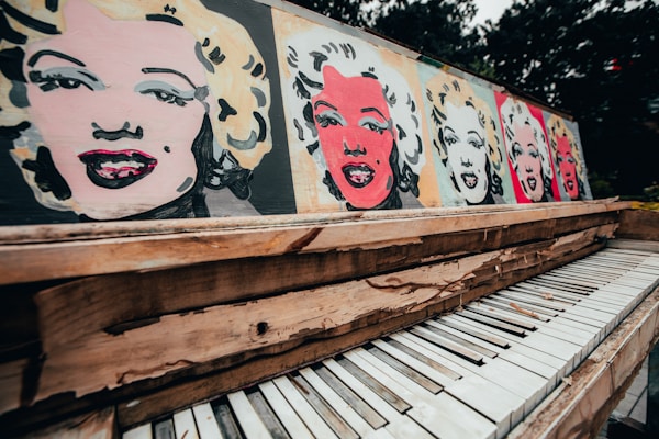 An artistic representation of famous pop art-style paintings featuring a woman's face on a wooden piano. The vibrant images are juxtaposed against the backdrop of an old, distressed piano with peeling paint and chipped keys. The setting appears outdoors with blurred greenery in the background.